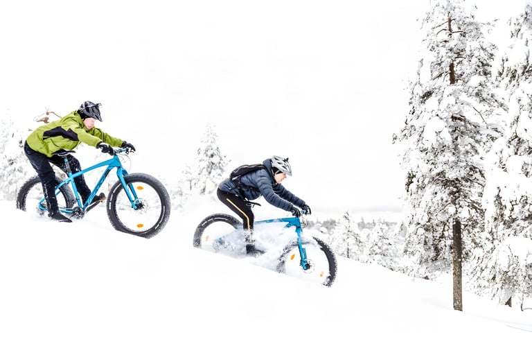 Johannes and Heikki of Roll Outdoors ride blue fatbikes down a snowy slope past frosted pines in Lapland.