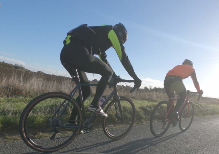 Two riders on a country road in low sun, one wearing the black Galibier Mistral jacket with hi-vis yellow panels.
