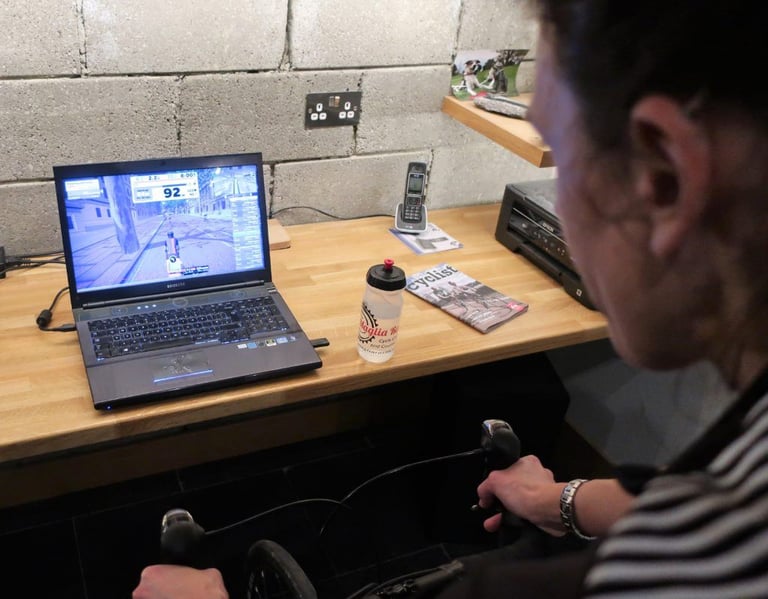 Rider's view of a Zwift ride on a laptop beside a water bottle, from the bars of a bike on a smart trainer.