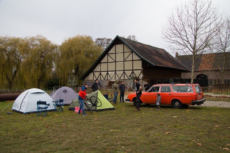 Tents pitched on grass beside a timber-framed barn, with a red classic Volvo estate, at the GravelFest campsite.