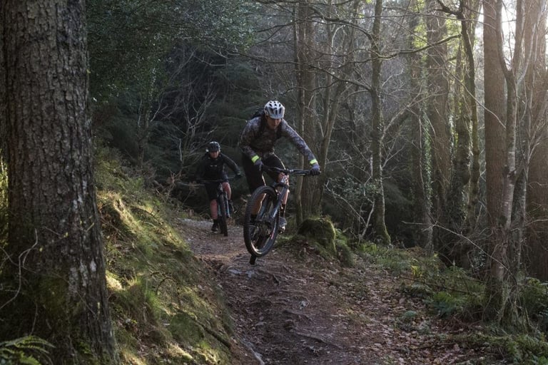 Wales360 rider lifts the front wheel on a steep woodland trail, with a second rider following behind.
