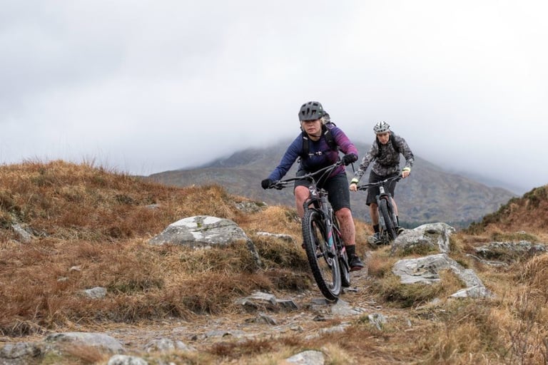 Two Wales360 riders on mountain bikes pick their way along a rocky upland trail under low cloud in Wales.