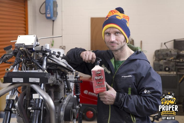 Guy Martin in a bobble hat holds a bottle of Proper Degreaser beside a motorcycle engine in a workshop.
