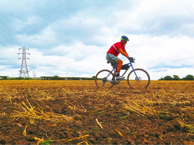 Rider on a flat-bar steel monstercross bike crosses a ploughed stubble field beneath electricity pylons.