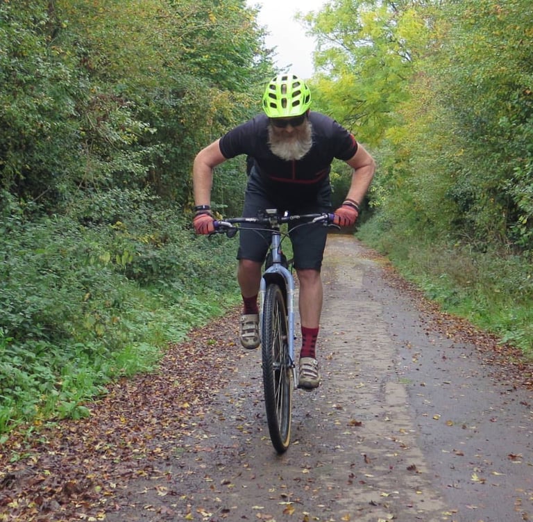 Bearded rider in a lime helmet rides a converted steel MTB up a leaf-strewn lane lined with autumn hedges.