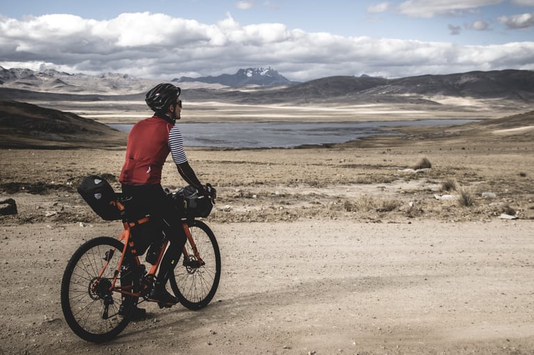 BikingMan founder Axel Carion pauses on a loaded orange bike on a dirt road above a lake, snow-capped peaks beyond.