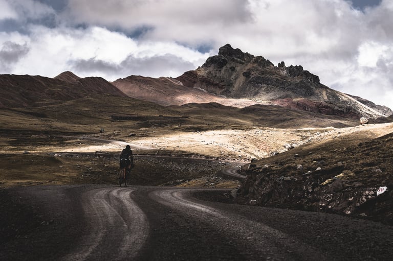 Lone cyclist on a winding gravel road below rugged red and grey mountain peaks under heavy cloud.
