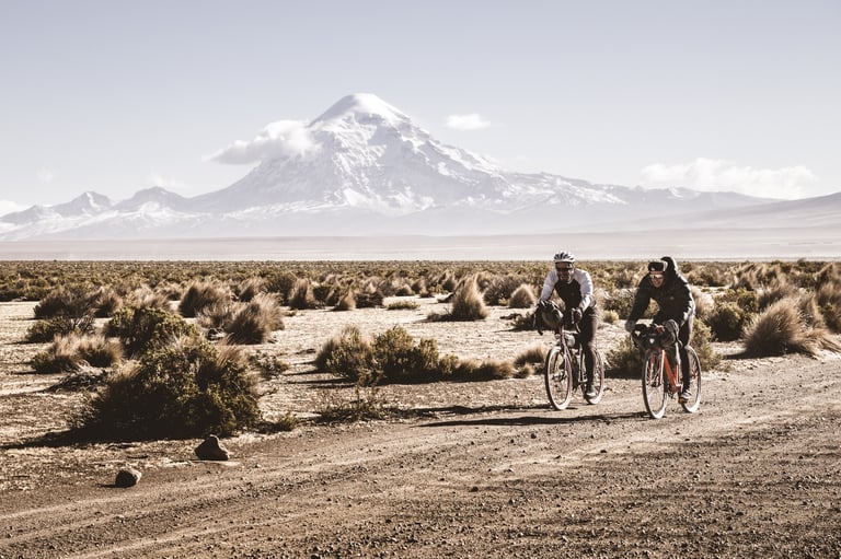 Two bikepackers ride a dusty track in Bolivia with a huge snow-covered volcano behind them.