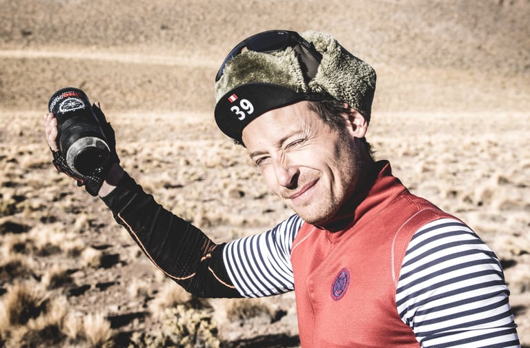 Axel Carion in a fur-trimmed cap with race number 39, grimacing as he holds up a near-empty bottle.