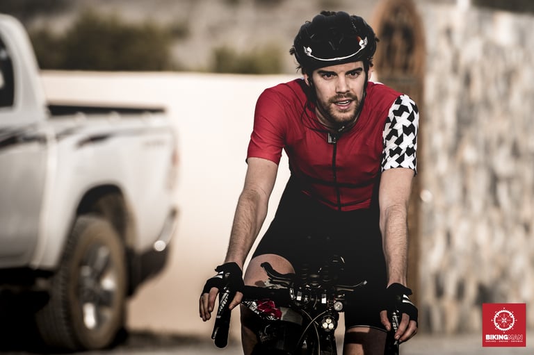Josh Ibbett in a red jersey rides towards the camera at BikingMan Oman 2018, a white pickup behind him.