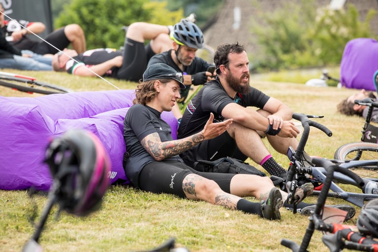 Riders relax on the grass beside purple inflatable loungers and their bikes at Grinduro Scotland.