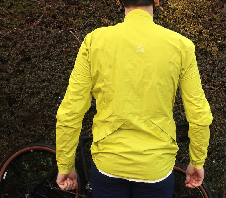 Rear of the 7Mesh Rebellion jacket showing the dropped hem and logo, worn in front of a hedge and bike.