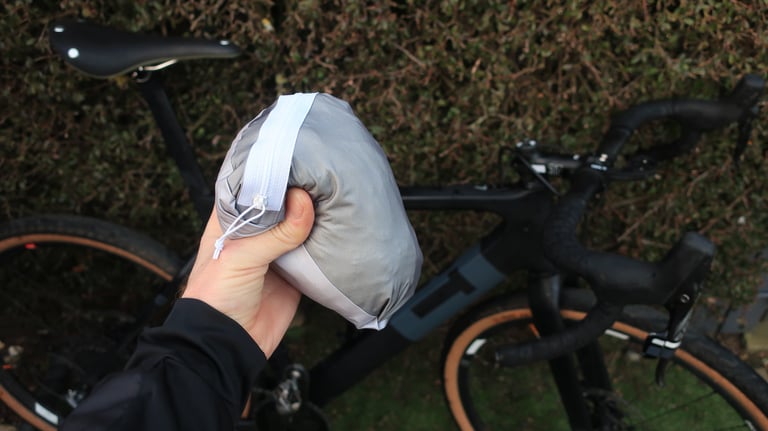7Mesh Rebellion jacket packed into its own grey pocket, held in one hand in front of a gravel bike.