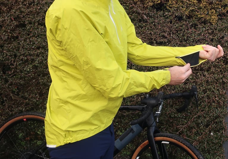 Rider unzipping the vented cuff of the 7Mesh Rebellion jacket to show the black mesh panel, gravel bike behind.