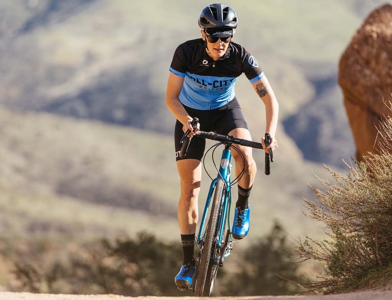 Head-on view of a rider climbing a dirt track on the teal Cosmic Stallion in a blue and black All-City Cycles jersey.
