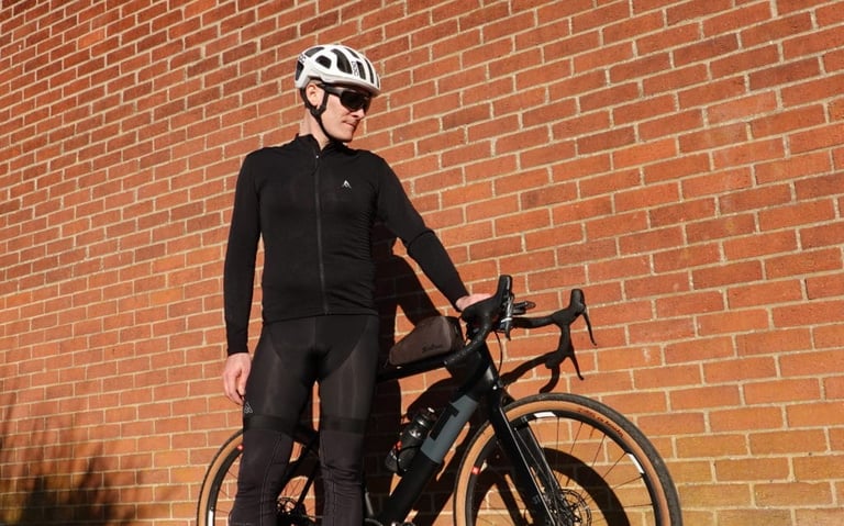 Rider posing against a brick wall in the all-black 7Mesh Mission Jersey and bib tights, with a gravel bike beside them.
