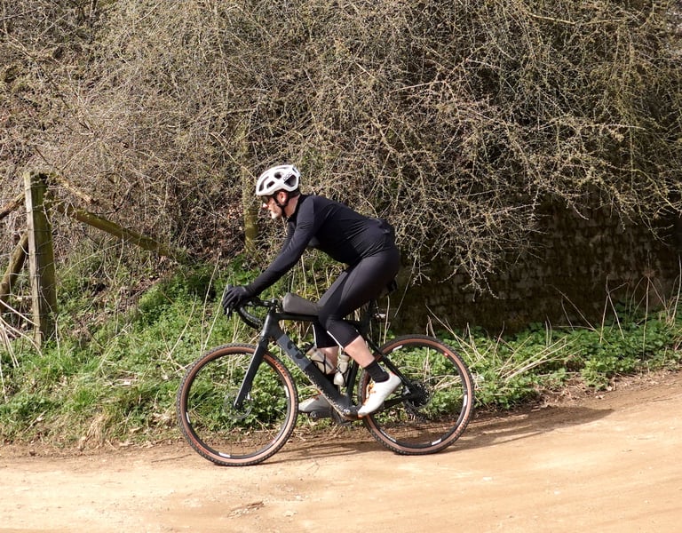 Rider in the 7Mesh Mission Jersey and black tights pedals a gravel bike along a dirt lane past a bare hedgerow.