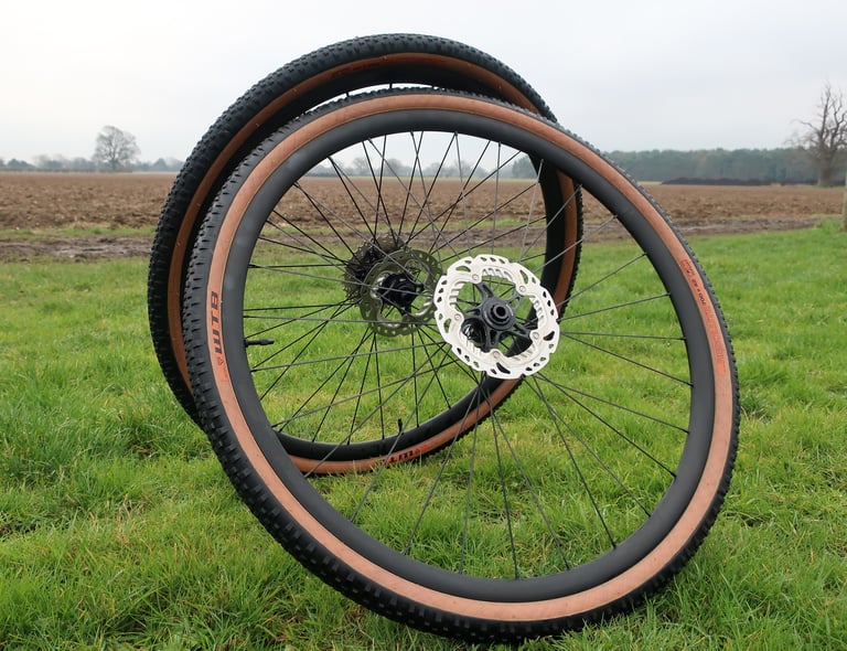 Sharp Precision Wheels gravel pair propped on grass, one wheel in front with disc rotor, ploughed field behind.