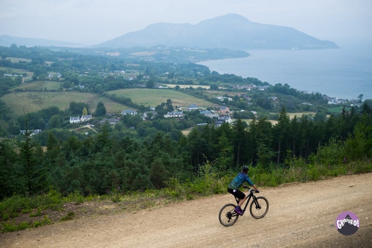 Grinduro rider on a gravel road above fields and the sea, with a hill across the bay, on the Isle of Arran.