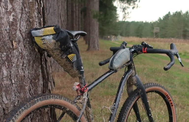 Apidura Racing Saddle Pack, mud-spattered, on a bike leaning against a pine tree beside a woodland track.