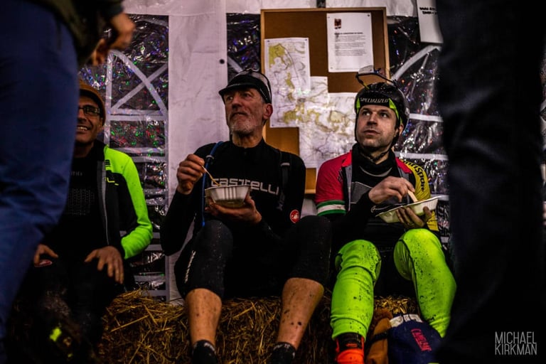 Riders eat from bowls on a hay bale in the finish tent at Yorkshire True Grit Dark Skies, with route maps behind.