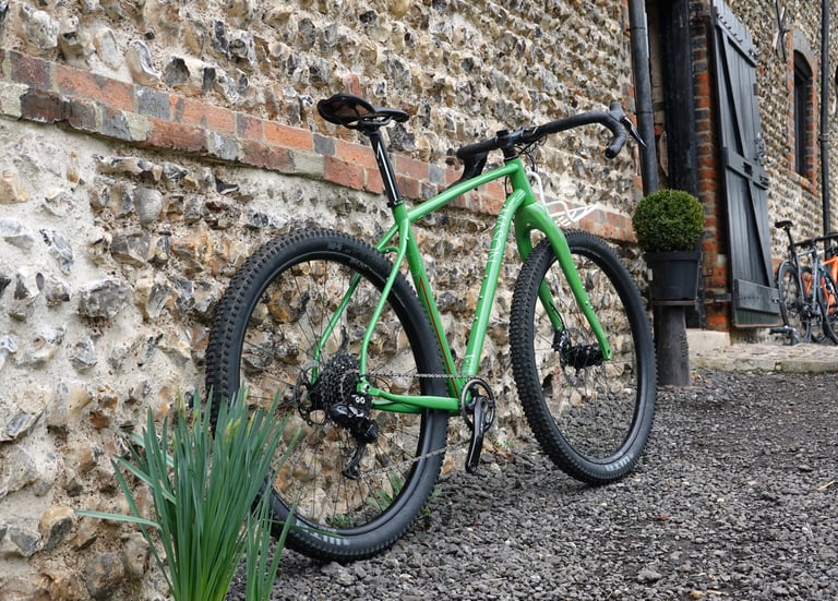 Green Mason InSearchOf adventure bike with big knobbly tyres leaning against a flint wall at the Mason Barn.