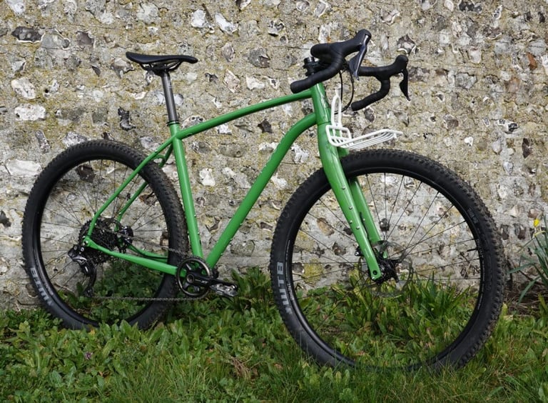 Side profile of the green Mason InSearchOf with drop bars, white front rack and knobbly 29er tyres against a flint wall.
