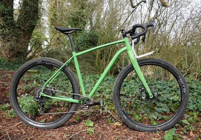 Mason InSearchOf in green propped among ivy and bare trees, showing its curved top tube and white front rack.