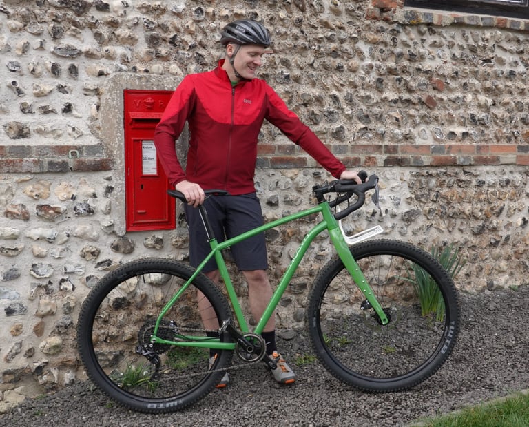 Rider in a red jacket stands beside the green Mason InSearchOf in front of a flint wall and red postbox.