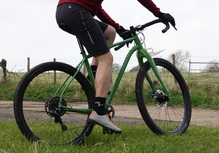 Rider's legs and the drivetrain of the green Mason InSearchOf, with knobbly tyres and a front rack, on a grassy track.