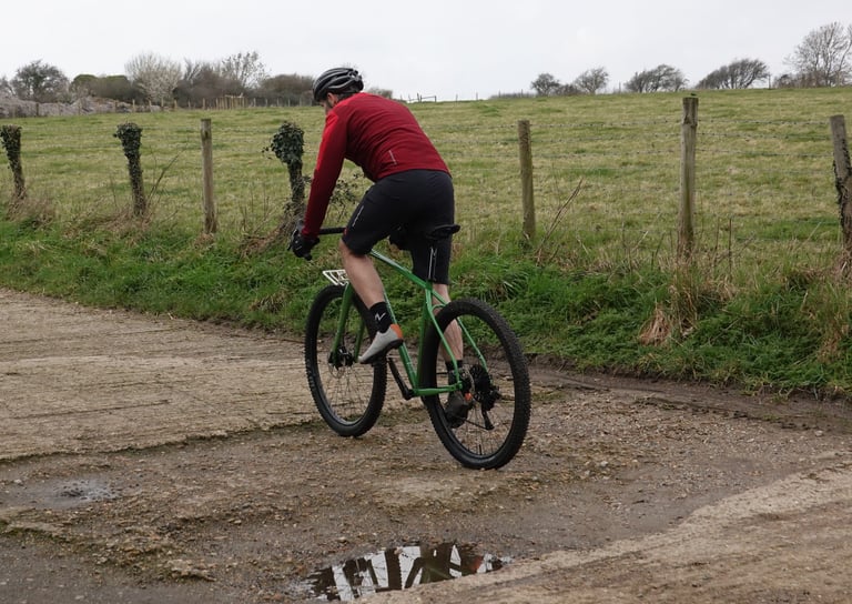 Rear view of a rider in a red jacket on the Mason InSearchOf crossing a muddy, puddled farm track.
