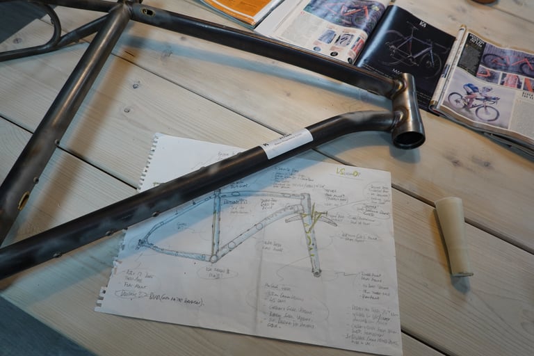 Hand-drawn InSearchOf design sketch beside an unpainted steel Mason ISO frame and bike magazines on a wooden table.