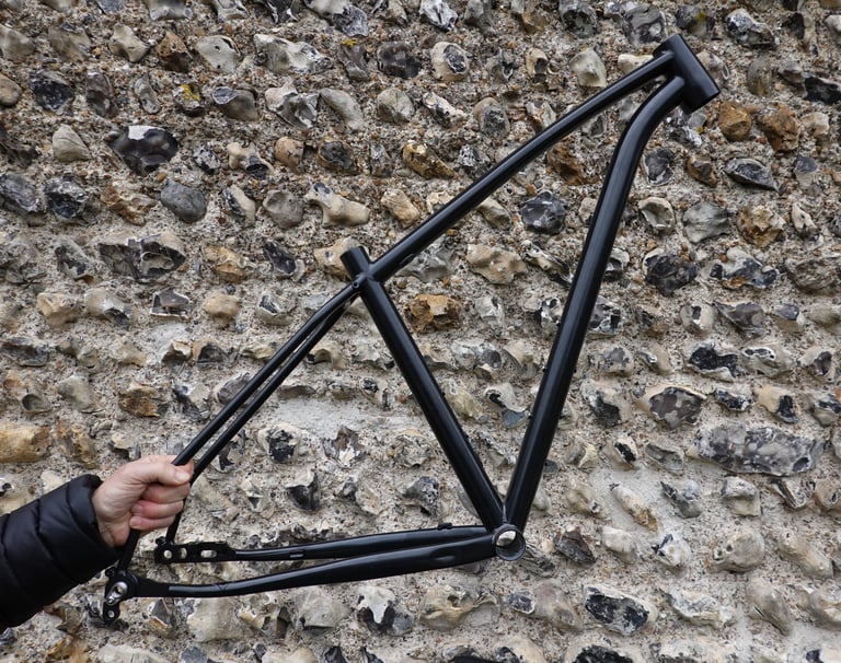 Bare black frame of the Mason ISO held against a flint wall, showing the triangle and its curved tubes.