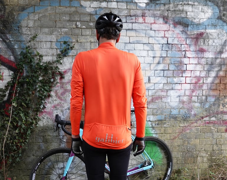 Back of the orange Galibier Roubaix Long Sleeve Jersey, showing the fit and rear pockets, with a bike behind the rider.