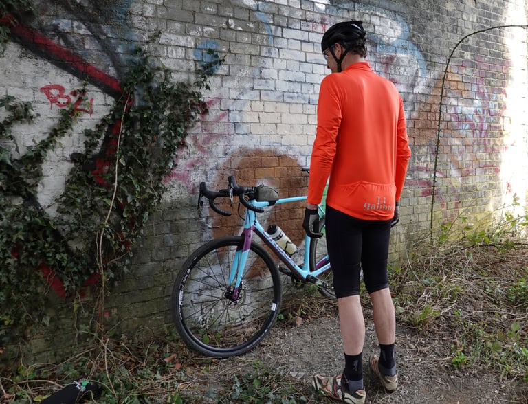Rider in the Very Orange Galibier jersey and black shorts stands beside a turquoise Orbea leant against a graffiti wall.