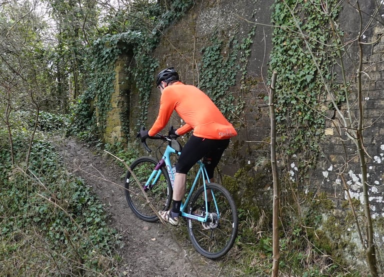 Galibier Roubaix jersey in Very Orange seen from behind as the rider pedals a rutted track beside an ivy-clad wall.