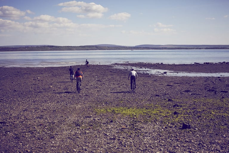 Four riders cross a stony shoreline beside calm water, with low hills beyond, at Brother in the Wild.