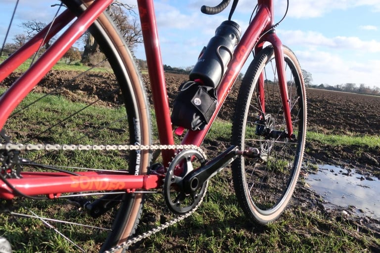 76 Projects Piggy on the down tube of a Sonder Camino AL gravel bike, with a muddy puddle beside the front wheel.