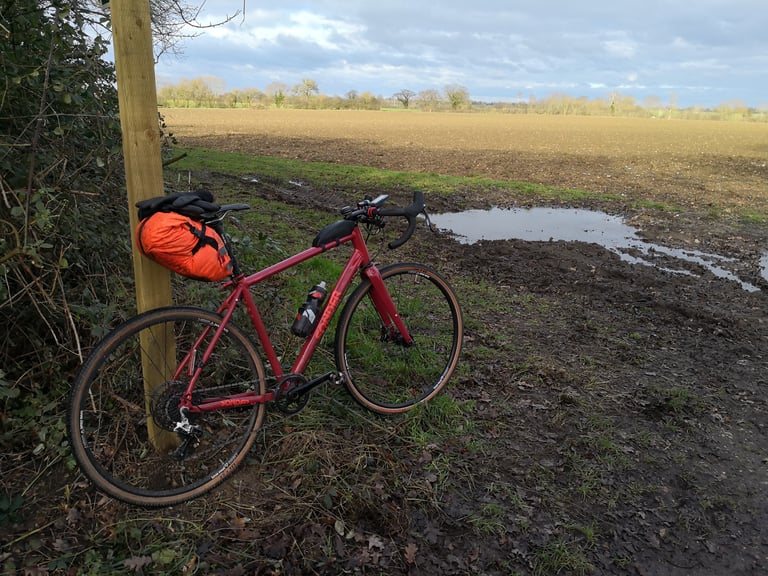 Sonder Camino AL propped against a fence post beside a muddy, puddled field edge, with an orange saddlebag.