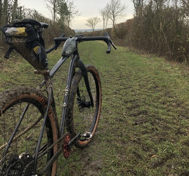Mud-covered Mason InSearchOf with a saddle pack and handlebar bag standing on a grassy byway.