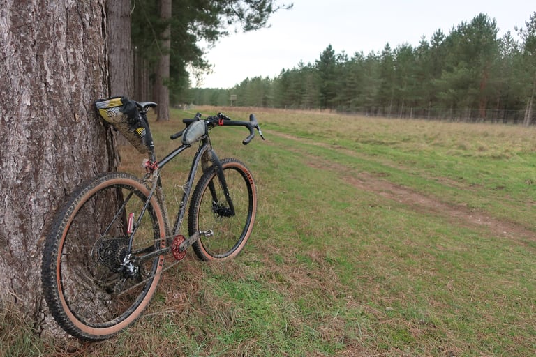 Muddy black Mason InSearchOf test bike with a saddle pack leaning against a pine tree beside a grassy track.
