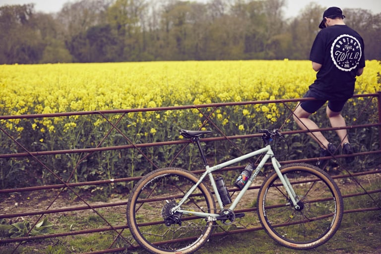Pale green Brother Cycles bike leaning on a rusty gate by a rapeseed field, with a rider in an event T-shirt perched on top.