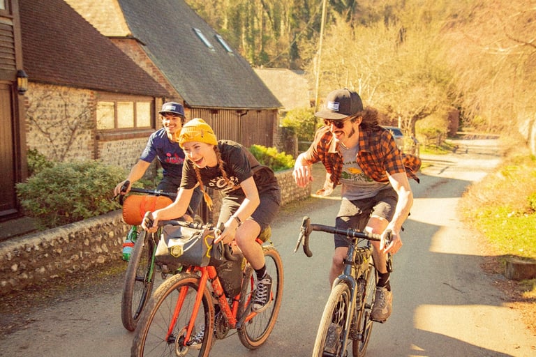 Three smiling riders in Morvélo Overland clothing ride Mason bikes along a village lane past flint cottages.