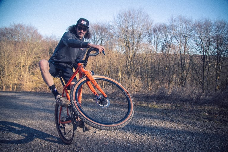Rider in sunglasses and Morvélo Overland clothing lifts the front wheel of an orange gravel bike on a gravel track.