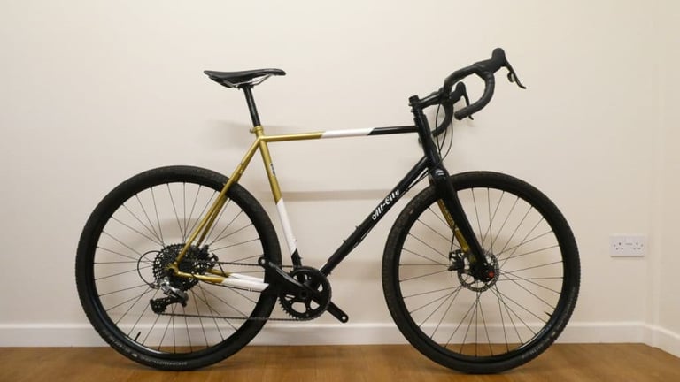 All City Cosmic Stallion steel all-road bike in gold, white and black, shown in side profile against a wall.