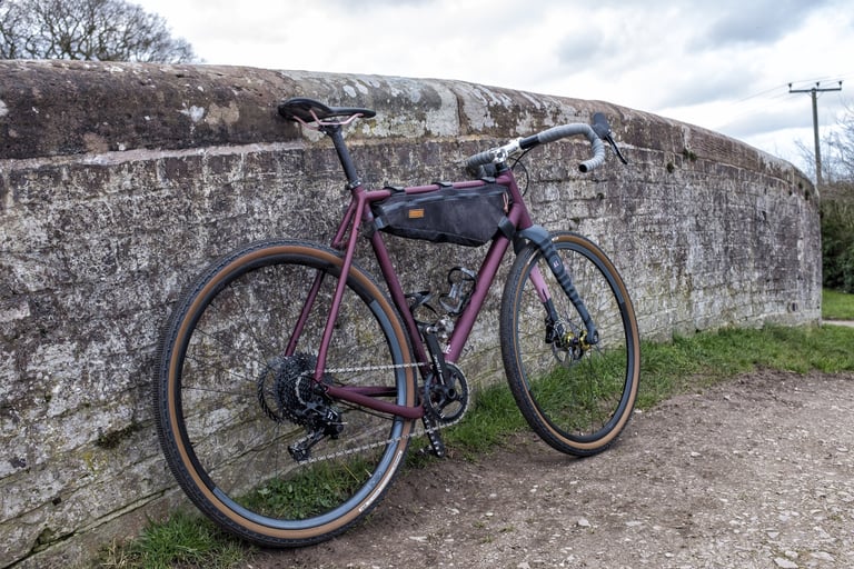 Rear three-quarter view of the Rondo Ruut AL on 650b wheels with a black frame bag, beside a stone wall.
