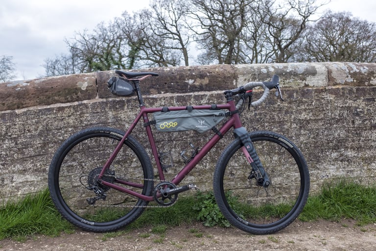Satin ruby Rondo Ruut AL with Apidura frame bag and Hunt wheels, side view against an old stone wall.