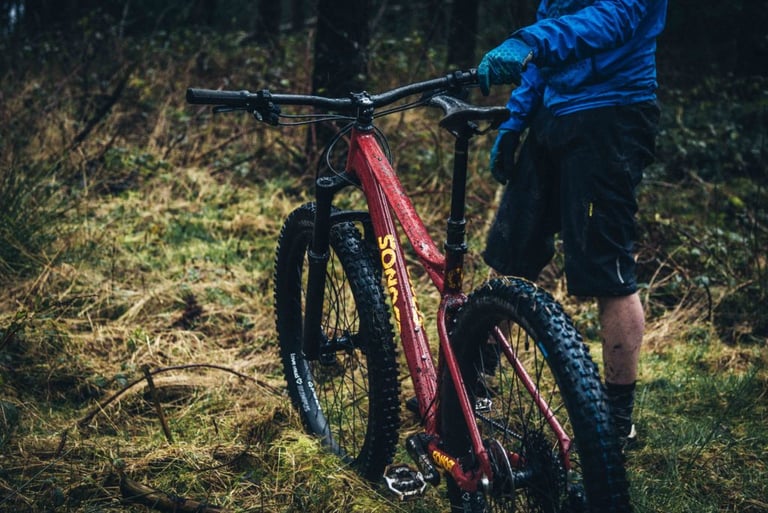 Muddy red Sonder Transmitter hardtail with chunky tyres and a rider in a blue jacket standing beside it in woodland.