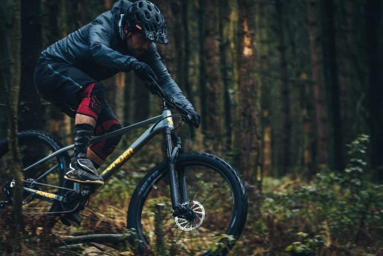 Rider in a dark jacket and knee pads corners hard on a grey Sonder Transmitter hardtail through woodland.