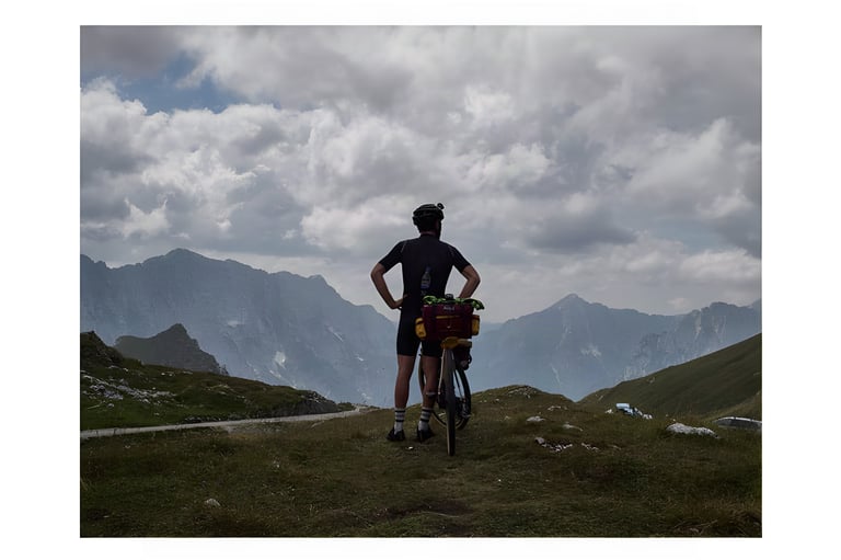 Rider stands with a loaded bike on a grassy pass, looking out over cloud-covered peaks in the Ariège.